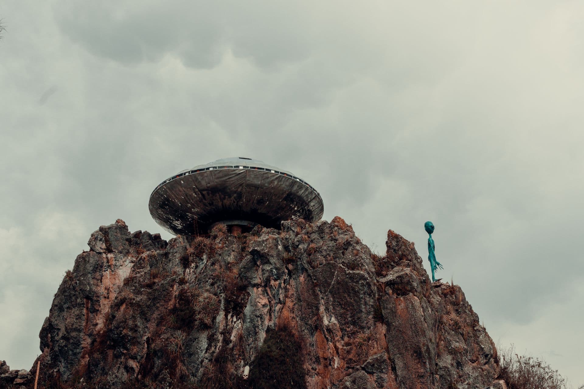 A silver disc-shaped craft resting on high ground beneath a heavy grey sky, with a thin dark figure standing nearby